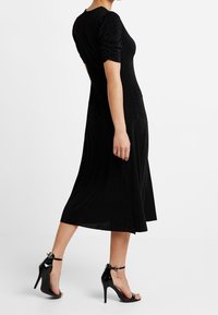 Sort kjole med rynkede korte ærmer, tætsiddende liv, og flared nederdel. Båret med sorte højhælede sandaler med en rem om anklen.