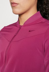 Primer plano de una persona que lleva una chaqueta deportiva magenta con cremallera, con un cuello acanalado y un pequeño logotipo oscuro de Nike en el pecho.