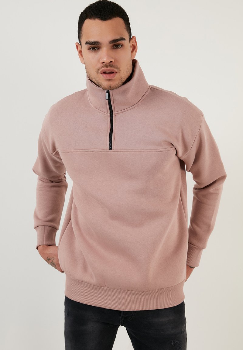 Suéter de pullover rosa claro con cremallera y cuello alto, con puños y dobladillo acanalados. Presenta una costura horizontal en el pecho. Textura suave.
