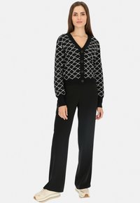 Czarny kardigan z białym geometrycznym wzorem, dekolt w serek, zapięcie na guziki, ściągacze przy rękawach i u dołu, w połączeniu z czarnymi spodniami i jasnymi sneakersami.