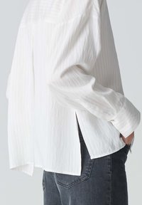 Weißes Langarmhemd mit vertikalen Streifen, weicher Textur, seitlichen Schlitzen und Manschetten. Kombiniert mit dunkelgrauen Jeans, die einen entspannten Schnitt zeigen.