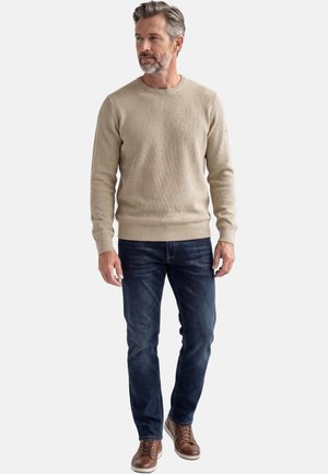 Mann mit grauem Haar und Bart, der einen beigen Pullover, dunkelblaue Jeans und braune Sneaker trägt, vor einem einfarbigen Hintergrund steht.