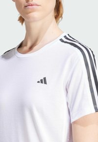 T-shirt de sport blanc avec un design à trois bandes noires sur les épaules, un tissu texturé et un petit logo noir sur la poitrine.