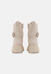 Botas de cuero beige con suela gruesa, acentos de hardware dorado y un interior texturizado. La parte posterior presenta dos correas y un detalle decorativo.