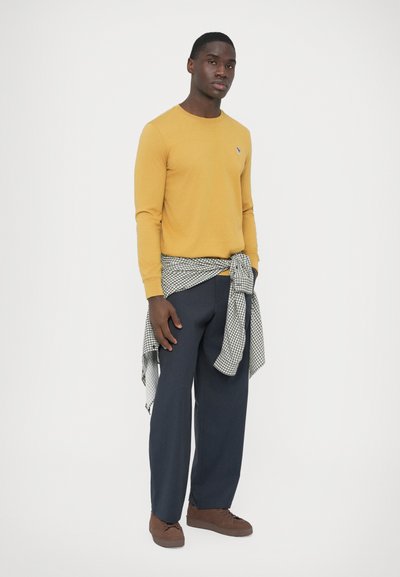 Pull jaune à manches longues avec petit emblème, pantalon large bleu marine, et chemise à carreaux noir et blanc nouée autour de la taille. Chaussures marron.