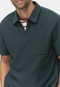 Mörkt tealfärgad kortärmad polo med dragkedja, med en strukturerad tyg och en liten bröstficka. Randig lager-lösning synlig under.