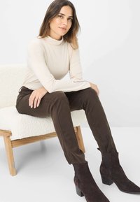 Beige langmouw turtleneck top, gecombineerd met donkerbruine aansluitende broek en donkere suède enkellaarzen, zittend op een gestructureerde witte stoel.
