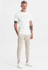 Witte t-shirt met "PALM ISLAND" logo, gecombineerd met lichtbeige sweatpants met geborduurde enkels en een trekkoord. Witte sneakers maken de outfit compleet.