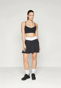 Fekete sportmelltartó fekete pliszírozott szoknyával, fehér derékkal. A modellen fekete cipő és fehér zokni van.