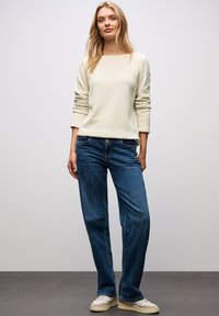 Helles beigefarbenes Langarmshirt, kombiniert mit dunkelblauen Jeans. Das Shirt hat einen quadratischen Ausschnitt und einen lockeren Schnitt, während die Jeans ein klassisches Design haben.