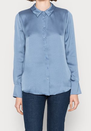 Frau trägt eine langärmelige, geknöpfte, hellblaue Satinbluse kombiniert mit dunkelblauen Jeans, steht vor einem einfarbigen Hintergrund.