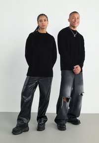 Două persoane poartă cămăși negre cu mâneci lungi. Una poartă pantaloni negri din piele lucioasă; cealaltă are blugi largi gri cu aspect uzat. Amândouă poartă pantofi negri.