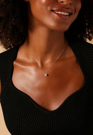 Gouden ketting met een kleine, heldere kristallen hanger. Close-up toont huidskleur, gestructureerde zwarte top en zachte halslijn.