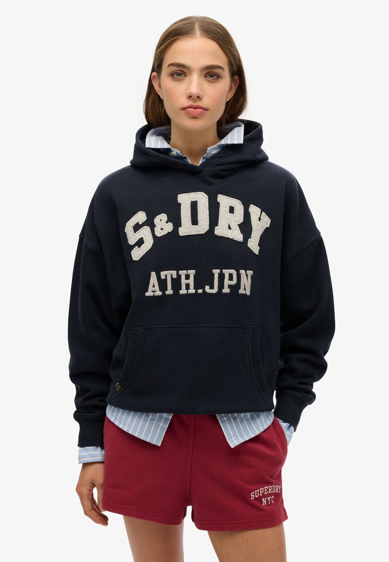 Marine hoodie met witte geborduurde tekst "S&D DRY ATH.JPN", kengaroozak, gedragen over een lichtblauw gestreept shirt en bordeauxrode shorts.