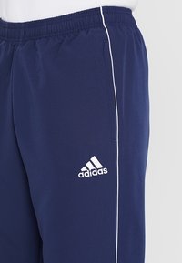 Mörkblå Adidas träningsoverallsbyxor med vit piping på sidan och elastisk midja, bärs med en vit topp.