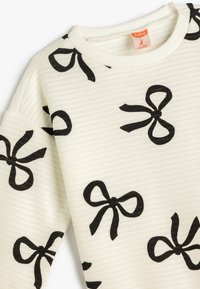 Sweatshirt crème à côtes avec motif de nœud noir, encolure ronde et coupe décontractée. Le tissu présente une texture subtile et des motifs contrastés.