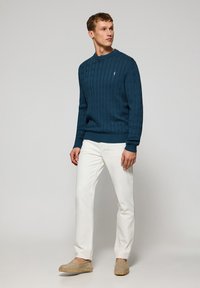 Kabeltrui in diep teal, met lange mouwen en ronde hals; gecombineerd met witte broeken; lichtbruine espadrilles met gevlochten detail.