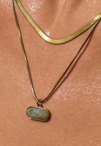 Halsband i gyllene metall med kedja som har en oval stenanheng med en ljusblå nyans och subtila fläckar, vilande mot huden med glans.