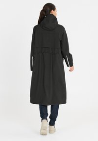 Zwarte lange jas met een capuchon, gesmokt rugdetail en lange mouwen. Gepaard met blauwe spijkerbroeken en beige laarzen. Gladde stoftextuur.