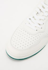 Hvite perforerte lær sneakers med hvite snører og en grønn gummisåle. Har en rund tå og glatte sømdetaljer.