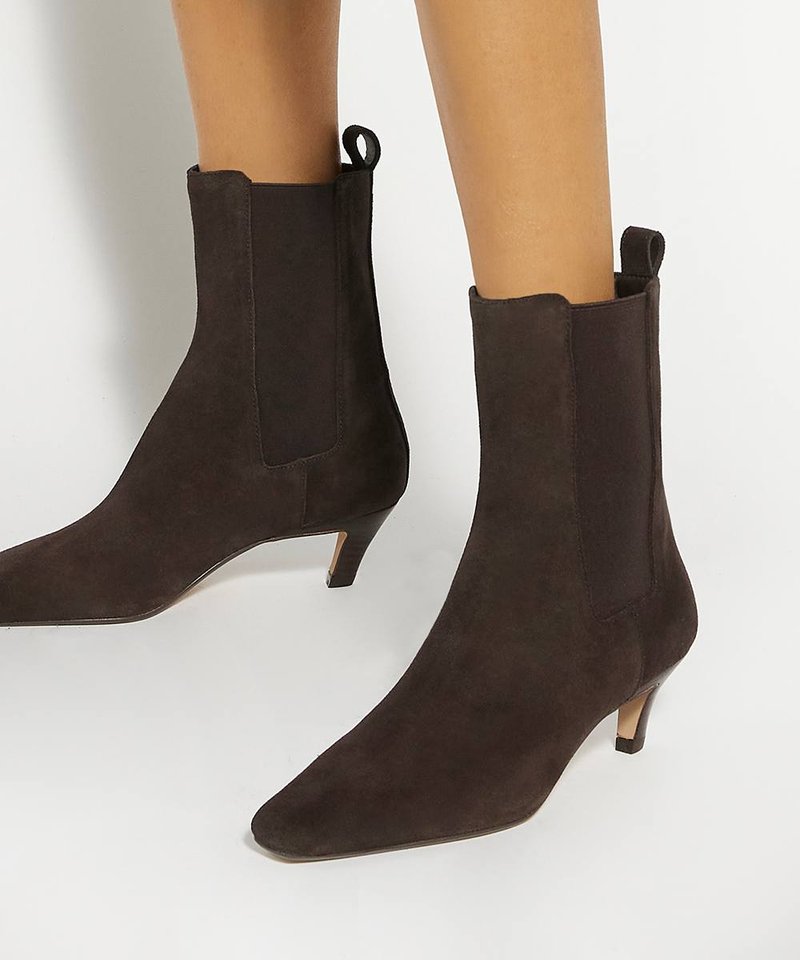 Bottines en daim brun foncé à bouts pointus et talons bas porte-mis sur jambes nues, sur fond blanc.