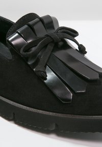 Mocassin en daim noir avec semelle épaisse, franges décoratives en cuir noir, et lacet en daim noué sur le cou-de-pied.