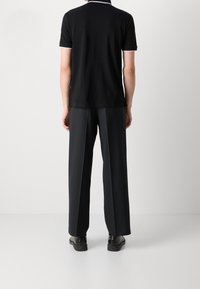 Svart kortärmad polo med vita detaljer på kragen och ärmsluten, i kombination med löst sittande svarta byxor och svarta skor, sedd bakifrån.