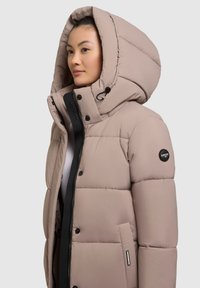 Helle beige Pufferjacke mit einer großen, strukturierten Kapuze, schwarzem Reißverschluss und Druckknöpfen; verfügt über ein Logo-Patch am linken Ärmel.