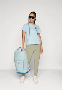 Šviesiai mėlynas kuprinė su žaliais akcentais, su priekine kišene, derinamas su šviesiai mėlyna marškinėlių ir kreminėmis kelnėmis.