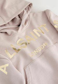 Světle růžová mikina z měkké látky se velkým zlatým nápisem "ALLSAINTS LONDON". Obsahuje přední kapsu a kapuci.