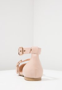 Zapato plano de cuero rosa claro con tira para el tobillo, que presenta herrajes en tono dorado y un back redondeado. Textura suave, diseño minimalista.