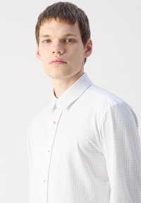 Camisa branca de botão com um subtil padrão geométrico azul, colarinho aberto, e textura de tecido leve, apresentando botões e um design ajustado.