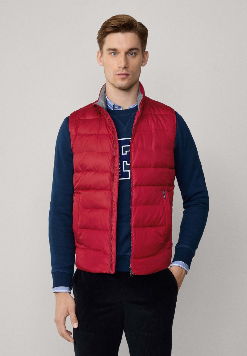 Chaleco acolchado rojo con un diseño de acolchado vertical, forro gris y cierre delantero. Lleva encima un sudadera navy con cuello redondo.