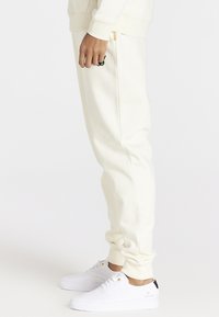 Krämfärgade sweatpants med avslappnad passform, försedda med elastiska muddar och en svart logotyp. Matchat med vita sneakers med guldiga detaljer.