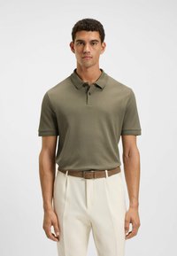 Olijfgroen polo t-shirt met korte mouwen en een twee-knops sluiting, gecombineerd met witte broek en een bruine leren riem. Gladde textuur.