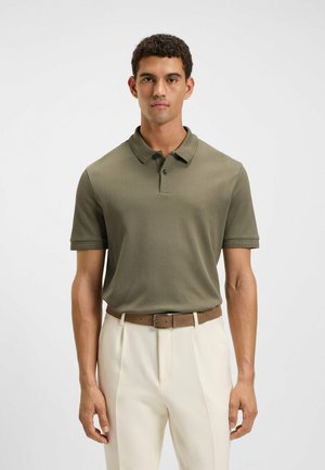 Olive green polo shirt with short sleeves and a two-button placket, paired with white trousers and a brown leather belt. Smooth texture.