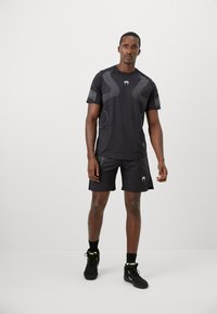 Sort atletisk skjorte med mesh-paneler, parret med sorte shorts. Har et logo og reflekterende detaljer for synlighed. Bæres med sorte sneakers.