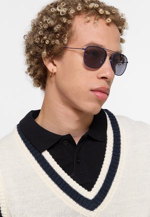 Giovane con capelli biondi ricci che indossa occhiali da sole scuri, un orecchino a cerchio dorato, una polo nera e un maglione bianco con righe marine.