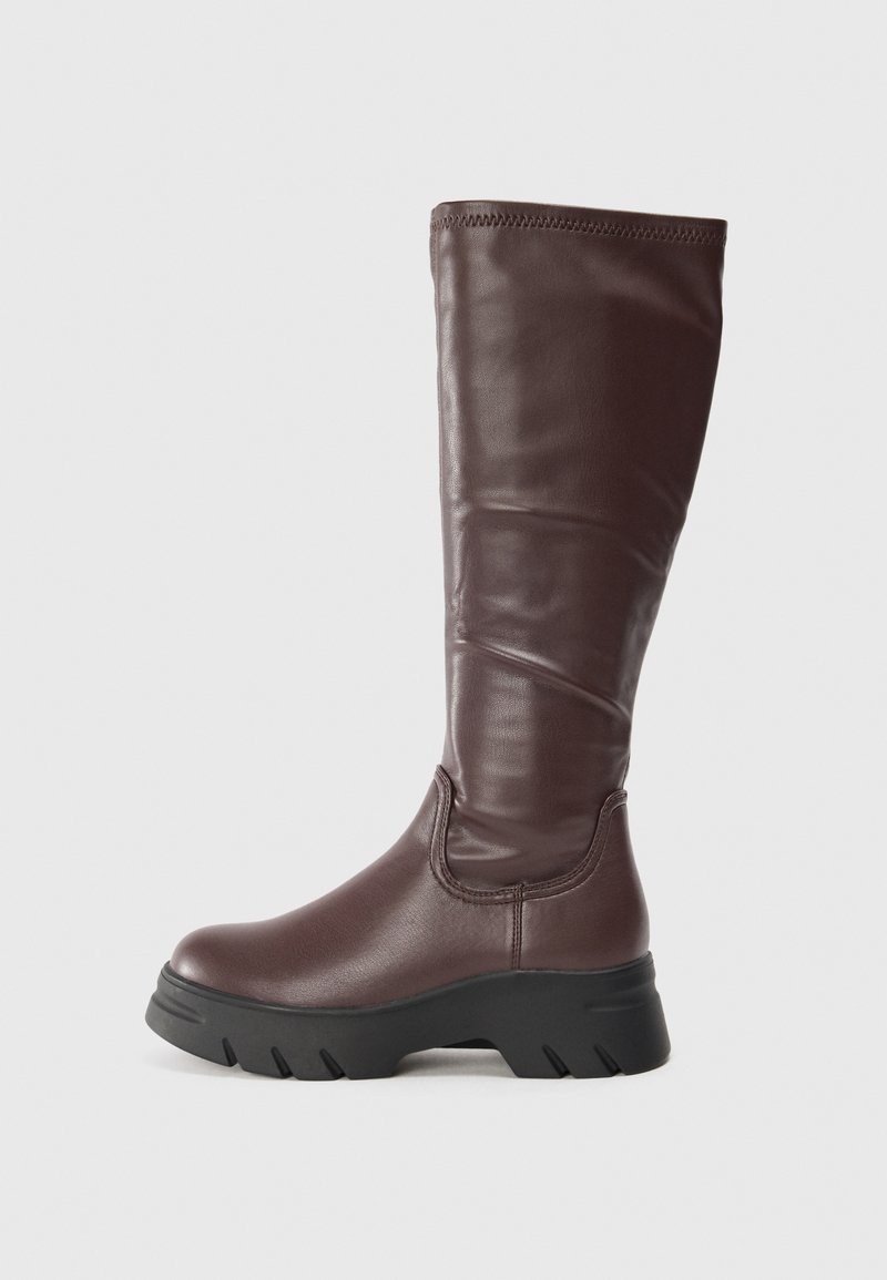 Bottes en cuir marron, montantes jusqu'au genou, avec une finition lisse, une épaisse semelle en caoutchouc noir et un minimum de détails de couture le long du bord supérieur.