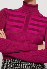 Pull à col roulé fuchsia avec texture côtelée et motif rayé horizontal. Fabriqué en matériau tricoté scintillant, comportant des manches longues.