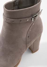 Šedá semišová kotníková bota s vysokým blokovým podpatkem, zdobená dekorativním páskem a kovovými nýty podél bočního švu pro zvýraznění detailu.