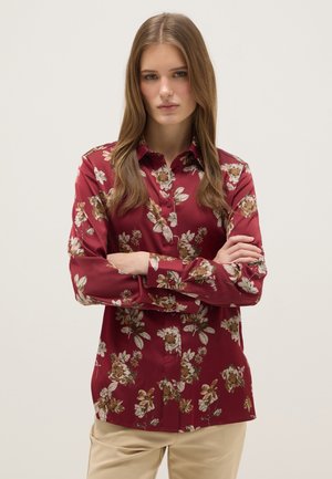 Mujer con cabello largo castaño claro, vestida con una camisa de botones con estampado floral en color burdeos y pantalones beige, de pie con los brazos cruzados frente a un fondo liso.