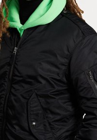 Svart isolerad bomberjacka med en slät yta, med dragkedja fram, sidofickor och en fastsydd ljusgrön huva under.