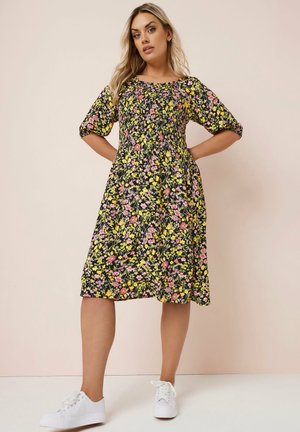 Robe noire à fleurs avec des manches courtes bouffantes, taille cintrée et une jupe à longueur genou ornée de motifs floraux jaunes et roses. Assortie à des baskets blanches.