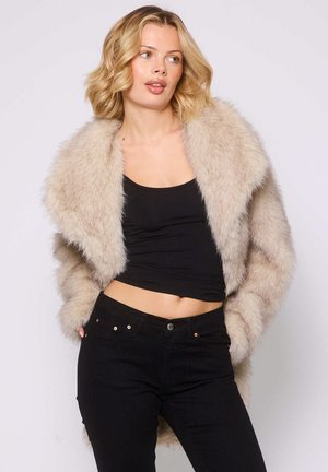Faux fur cropped jakke i lys beige, med en oversized krave og lange ærmer, parret med en sort tætsiddende top og jeans.
