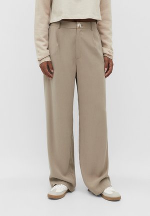 Person iført løstsiddende beige bukser, hvide sneakers og en lysebeige langærmet top, der står mod en ensfarvet baggrund.