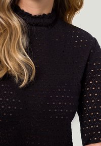 Sort top med en høj, flæset hals og et tekstureret mønster af små prikker og åbninger i stoffet, korte ærmer.