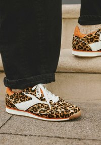 Leopardenmuster-Sneaker mit orangen Akzenten, weißen Lederdetails, weißen Schnürsenkeln und einer strukturierten Oberseite. Schwarze Jeans mit sichtbaren Umschlägen.