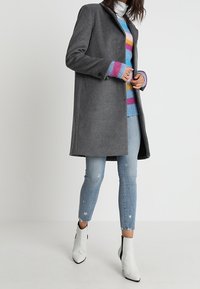 Šedý vlněný kabát s nařezaným střihem, nošený přes barevný pruhovaný svetr. Kombinován se světlými modrými džíny s hvězdnými vzory a bílými kotníkovými botami.