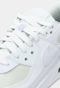 White and light gray Nike sneaker close-up showing textured mesh, leather panels, lace eyelets, and thick white laces tied across the tongue.